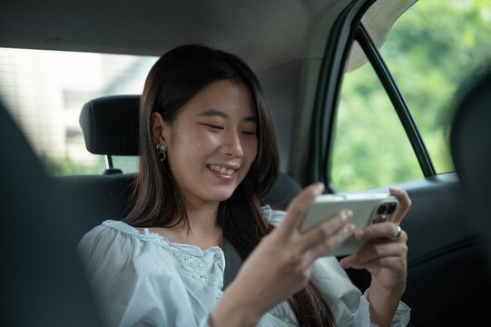 Young Asian woman watching clip on smartphone while riding comfortably in back seat of taxi cab car.