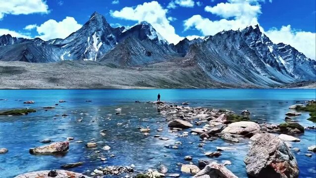 Person standing with arms raised on rocky lake shore surrounded by clear blue water and snow capped mountain peaks under sunny sky
