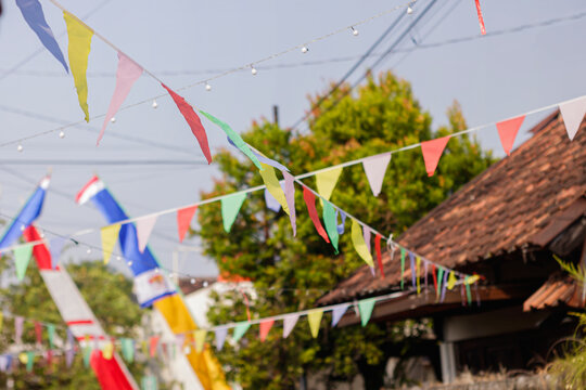 Close focus on multicolored triangular pennant flags hanging outdoors with a traditional village house roof and lush green trees in the background. High Quality Photo