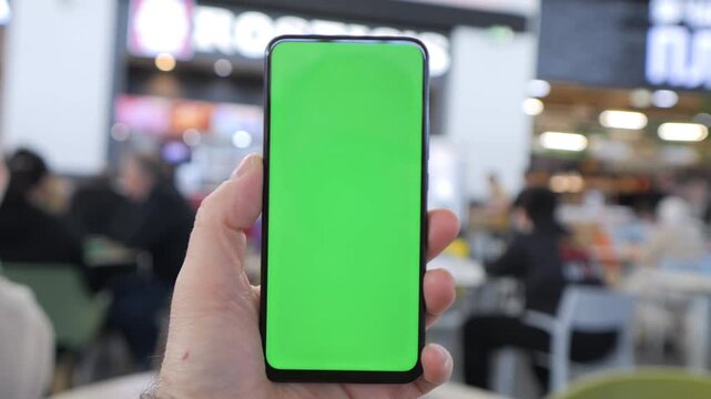 Hand holds smartphone with green screen. Background shows busy indoor space. People sit and walk around casually. Ideal for app demos or digital content. Realistic setting with natural lighting