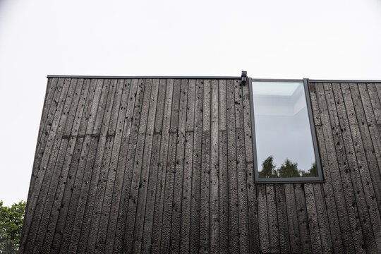 Low-angle view of a modern facade clad in black Shou Sugi Ban timber, featuring vertical charred boards, a tall reflective window, trees, and ample sky copy space