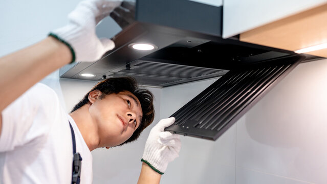 Asian male technician removing range hood filter looking at exhaust fan inside the cooker hood. Maintenance worker man wearing gloves repairing modern kitchen hood. Home appliance service on site