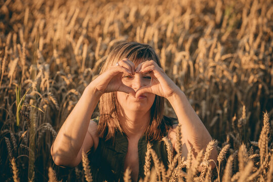 Female model making heart shape with hands while sitting in a golden wheat field during sunset, surrounded by tall wheat stalks and warm sunlight
