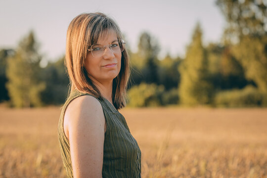 Woman with glasses standing in a golden field during sunset, wearing a green sleeveless top, with trees and a clear sky visible in the background