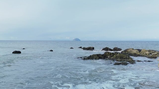Waves break over rocks on Ayrshire coast with Ailsa Craig visible on horizon.