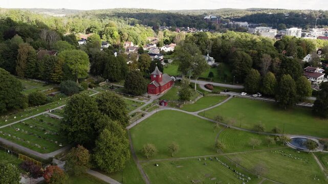 Aerial drone view of Rada Church surrounded by parkland, mature trees and walking paths. Historic red wooden church stands within landscaped grounds near Molnlycke in Harryda, Sweden
