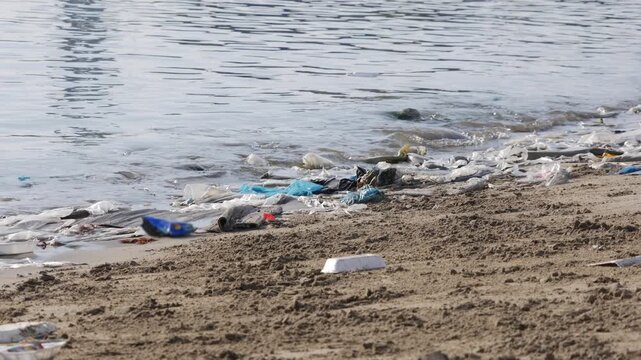 Plastic trash and discarded packaging scattered along a sandy beach by the water. Polluted seascape.