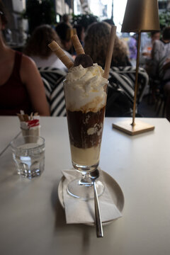 Elegante coppa di gelato affogato al caff&egrave; con strati di crema, cioccolato, panna montata e cialde croccanti, servita su un tavolo da bar all'aperto con lampada di design e atmosfera estiva.