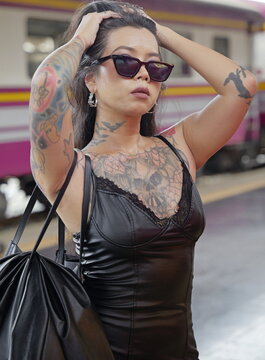 Young stylish heavily tattooed Asian woman with long dark hair and sunglasses poses on a train platform with a passenger train in the background