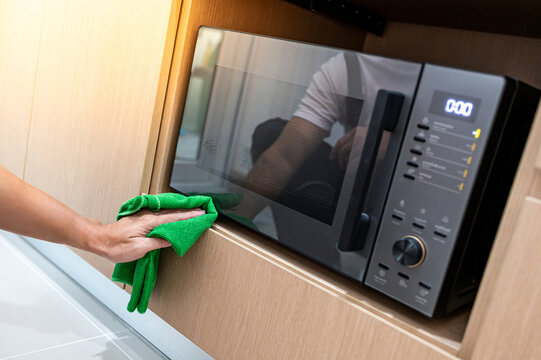 Male hand holding green microfiber cloth wiping on modern black microwave oven in wooden cabinet. Cleaning home appliance in the kitchen