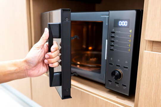 Hand holding door handle opening black microwave oven in modern wooden cabinet. Home appliance in the kitchen