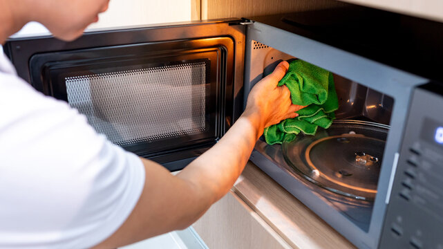 Male hand holding green microfiber cloth wiping inside the black microwave oven in wooden cabinet. Cleaning home appliance in the kitchen