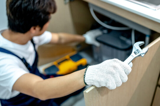 Asian male plumber with glove and adjustable wrench in his hand opening wooden cabinet checking grease trap box and pipe system under the kitchen sink. Handyman fixing plumbing system in the kitchen.