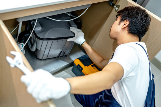 Asian male plumber with glove and adjustable wrench in his hand opening wooden cabinet checking grease trap box and pipe system under the kitchen sink. Handyman fixing plumbing system in the kitchen.