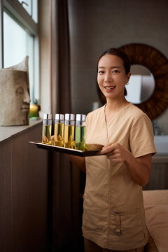 Happy Korean therapist using natural massage oils for wellness spa treatment and looking at camera.