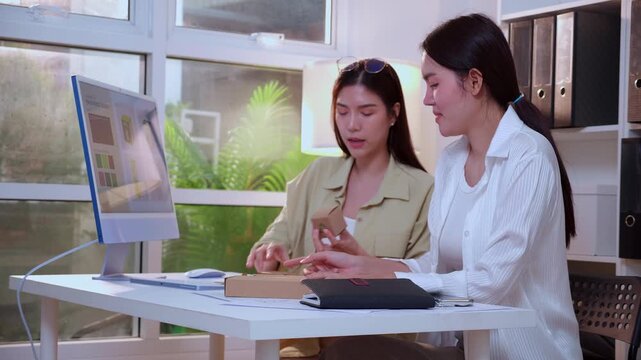 Two young asian woman designer working on packaging design with ai software with desktop computer, business team discussion of eco friendly cardboard box product development at modern home office.