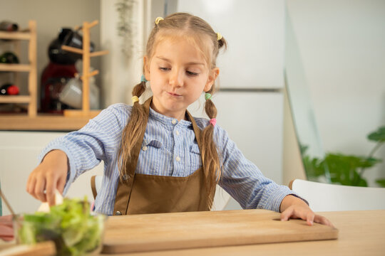 Cute little girl pressing a bread slice on wooden cutting board while making sandwich in kitchen. Adorable caucasian child enjoying preparing healthy breakfast snack at home kitchen table.