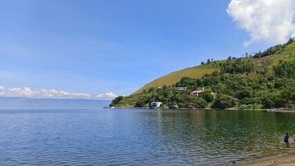 Fototapeta premium Scenic view of Lake Toba shoreline with green hills, houses, and boats under blue sky. Calm water reflects the landscape.
