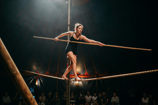 A female tightrope walker in a black leotard balancing on a rope with a long wooden pole inside a dark circus tent with an audience watching