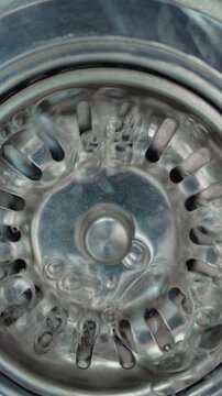 Water Flowing Into Sink Drain. Clear water swirls across a stainless steel basin and drains through a metal strainer.