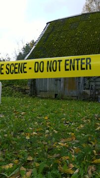 Yellow crime scene tape with the words 'do not enter' blowing in the wind in front of an old abandoned rural farmhouse and garden on a cloudy autumn day, cordoning off the area