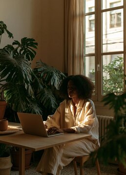 Young woman sitting at a wooden table in front of a large window. she is wearing a white blouse and has curly hair. the woman is working on her laptop and appears to be focused on her work.