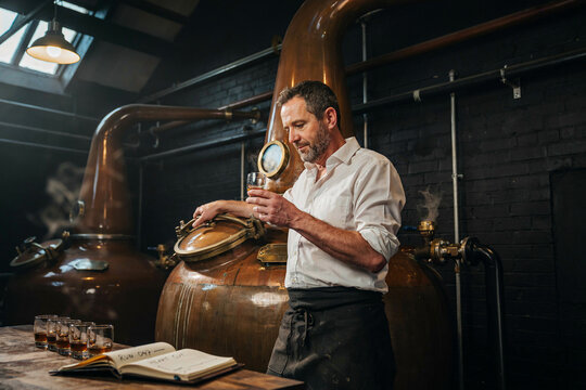A man in a white shirt and dark apron smelling a glass of spirit while standing in a distillery with large copper stills and brick walls