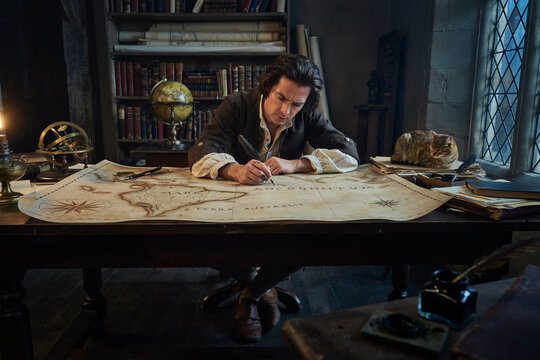 A man in historical clothing sitting at a desk in a library on an old map with a quill pen while a cat sleeps on a stack of books