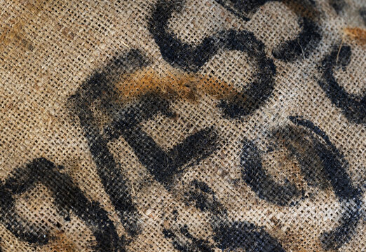 Worn burlap coffee sack close-up showing coarse jute weave, black stencil lettering and stains. Rustic textile texture ideal for backgrounds, packaging, agriculture, vintage and industrial design