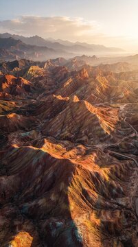 Aerial sunrise view of colorful sandstone hills in Zhangye, China, featuring vivid mineral layers, dramatic shadows, distant mountains, and intricate geological formations.