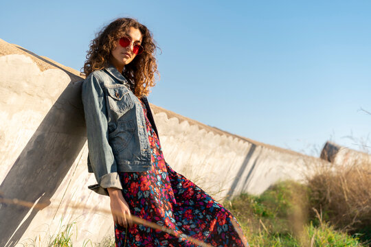 Portrait of young woman with curly brown hair wearing red sunglasses leaning against wall