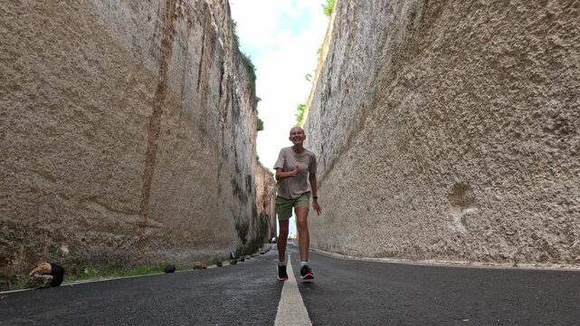A happy young blonde woman runs and jumps energetically on a narrow road between two tall cliffs leading to the sea on Bali island during daytime.