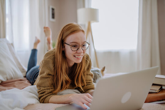 Smiling woman wearing eyeglasses using laptop lying on bed at home