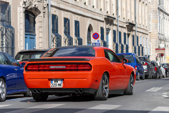 Nancy, France - March 30th 2019 : View on an orange Dodge Challenger driving in a street with parked cars and buildings on the side.