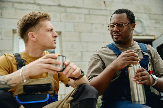 Caucasian young adult man and African American man wearing overalls, talking during coffee break at construction site, representing teamwork in home renovation, medium shot