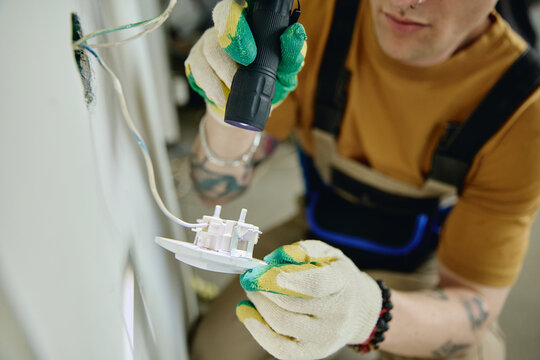 Young adult Caucasian electrician installing power outlet during house renovation, holding flashlight. Image suitable for home improvement services and electrical repair marketing