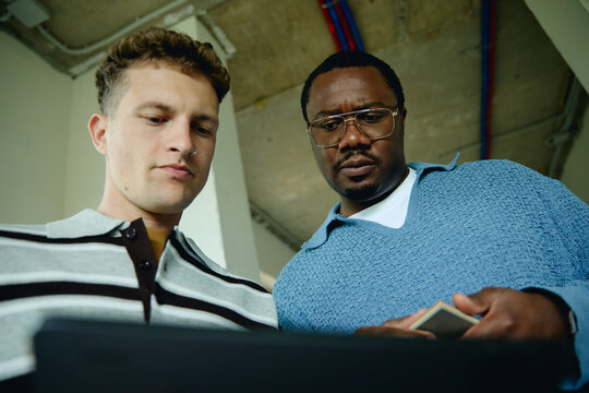 Diverse young adult Caucasian man and African American man inspecting house renovation plans on digital tablet. Low angle shot showing teamwork in construction and real estate