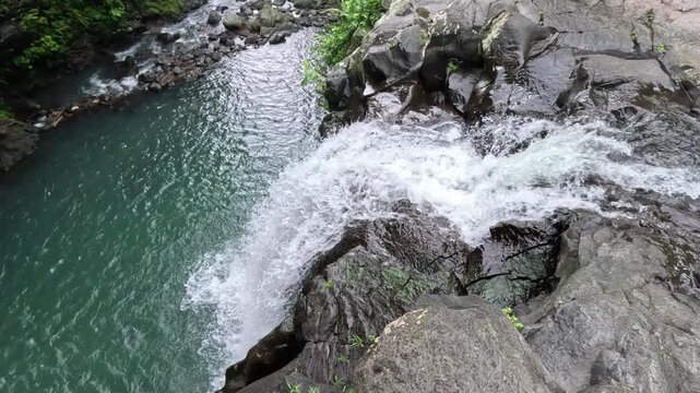 Aerial view of a waterfall flowing over rocks surrounded by lush greenery on Bali island. The waterfall leads to a natural swimming river on a cloudy day in Ubud.