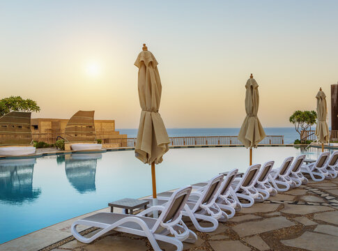 Luxury infinity pool at coastal resort during golden sunset, featuring closed umbrellas and empty lounge chairs overlooking serene blue ocean. Concept - peaceful high-end vacation, wellness travel.