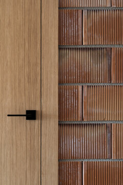 Closeup wooden door with matte black handle beside textured brown brick wall showing vertical ribbed pattern and warm natural tones, combining smooth wood grain, masonry detail, and modern interior