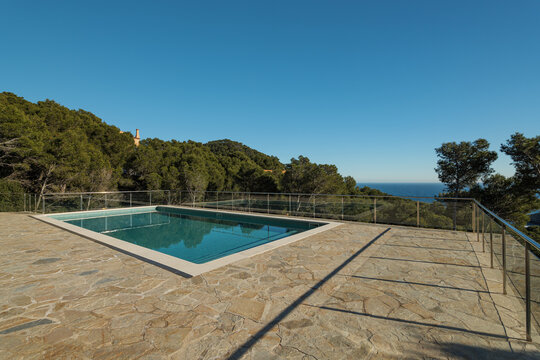 Modern outdoor swimming pool with stone terrace, lush greenery, clear blue water, and distant ocean horizon under bright sunlight in upscale residential setting