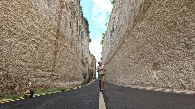 A young blonde woman smiles while walking along a road between two white cliffs on Bali island, capturing photos of the unique rock formations on her phone.