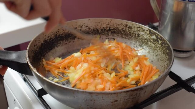 Hand-held slotted metal spatula stirs diced onion and julienned carrot in worn frying pan on gas stovetop, showing home cooking, ingredient preparation, and rustic kitchen texture, with onion, carrot