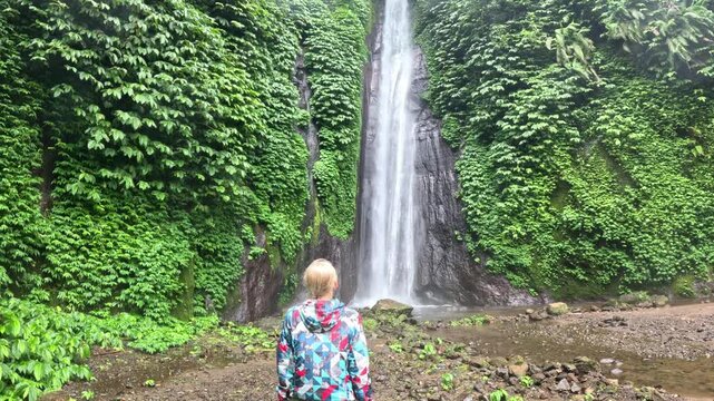 A young blonde woman in a colorful raincoat and green shorts stops to admire a large waterfall surrounded by lush plants on a rainy day on Bali island.