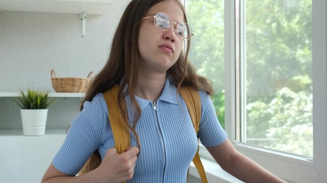 Pensive unhappy teen schoolgirl in glasses puts on yellow backpack at home looking out of window. Back to school again concept