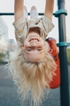 A girl with curly blond hair hangs upside down on a horizontal bar in Dovre in the summer. A happy childish look and long hair.