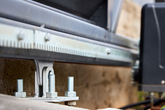 Sliding gate motor mechanism with metal gear rack, drive pinion, heavy duty foundation anchor bolts, steel support bracket mounted on concrete outdoor structure for automatic entrance system.