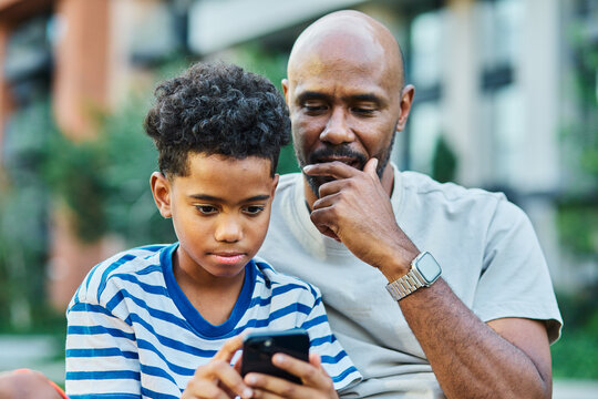 Portrait of father and son having fun using a mobile phone outdoors in park or nature,, family life, parenting, love and bonding concepts