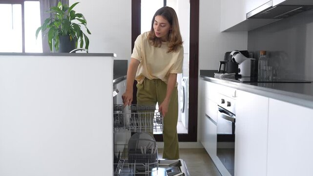 Woman loads dirty dishes into dishwasher in a modern kitchen. Housewife cleans a bright kitchen, disassembling the dishwasher. Concept of cleanliness, cleaning, and technology.