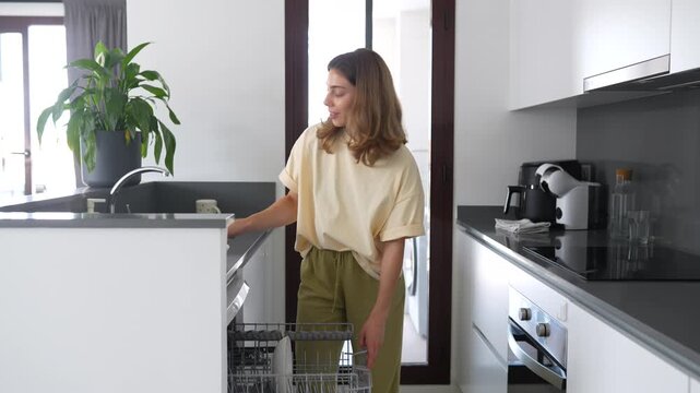 Woman loads dirty dishes into dishwasher in a modern kitchen. Housewife cleans a bright kitchen, disassembling the dishwasher. Concept of cleanliness, cleaning, and technology.